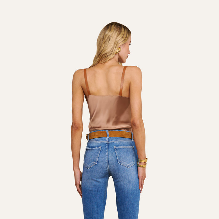 A woman stands facing away, wearing the Lux Silk Buckle-Strap Camisole in brown with vegan leather straps, paired with blue jeans, a brown belt, and gold bracelets. Her blonde hair is down as she looks slightly to the left against a plain off-white background.