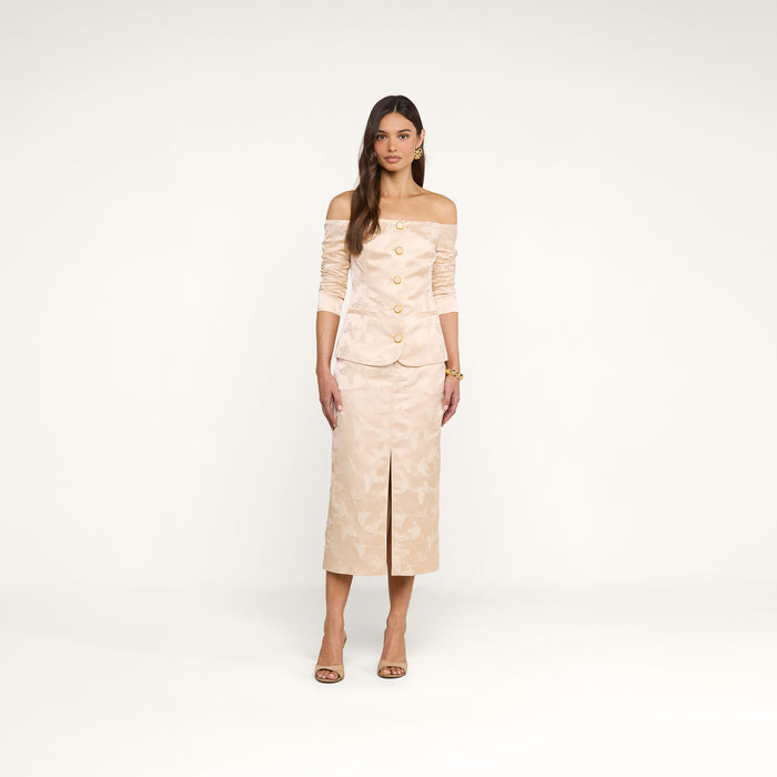 A woman stands against a plain white background wearing the Alice Blazer Top, a cream-colored, off-the-shoulder blazer with gold buttons, paired with a matching below-the-knee skirt featuring a front slit and heels.