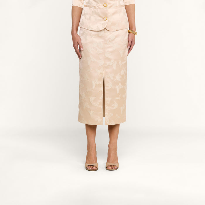 A woman stands facing forward against a plain white background, wearing the Madonna Midi Skirt with a beige butterfly jacquard floral pattern as part of a matching skirt suit. Her hands rest relaxed at her sides, and she accessorizes with open-toed heels and a bracelet.