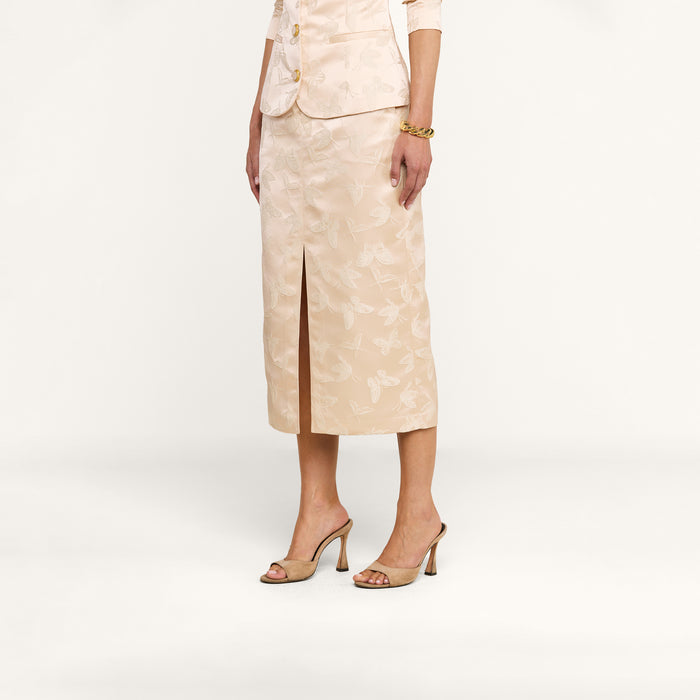 A woman stands against a plain white background, arms at her sides, wearing the Madonna Midi Skirt—a light beige skirt in butterfly jacquard with a floral pattern and a front slit—paired with high-heeled sandals.