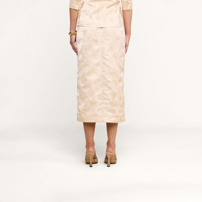A woman stands facing away, wearing the Madonna Midi Skirt in light beige with a matching top featuring elbow-length sleeves. She pairs the outfit with high-heeled shoes against a plain white background.