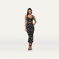 A woman stands against a plain, light background wearing a black, sleeveless crop top paired with the Mae Knit Flare Skirt—a high-waisted, polka dot fit and flare skirt—and black sandals.
