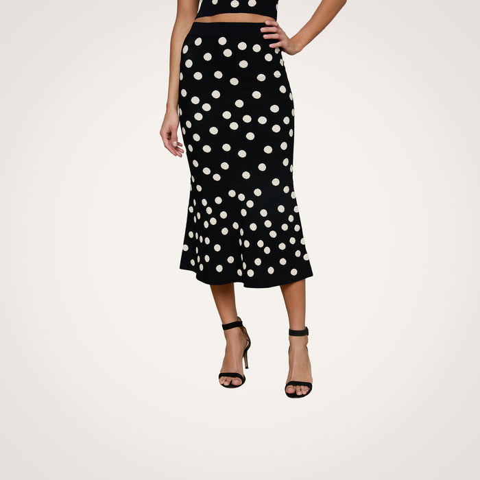 A woman wearing the Mae Knit Flare Skirt—featuring bold white polka dots on black—paired with a black sleeveless top and black high-heeled sandals, stands against a plain light background.