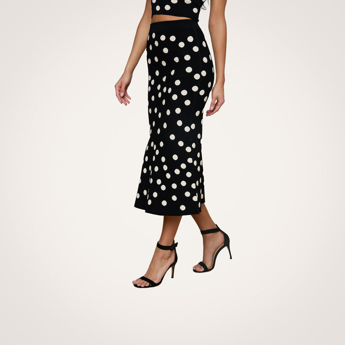 A woman wears the Mae Knit Flare Skirt in black polka dot, paired with coordinating high-heeled sandals and a matching polka dot top. The fit and flare design of the skirt creates a timeless silhouette against the plain white background.