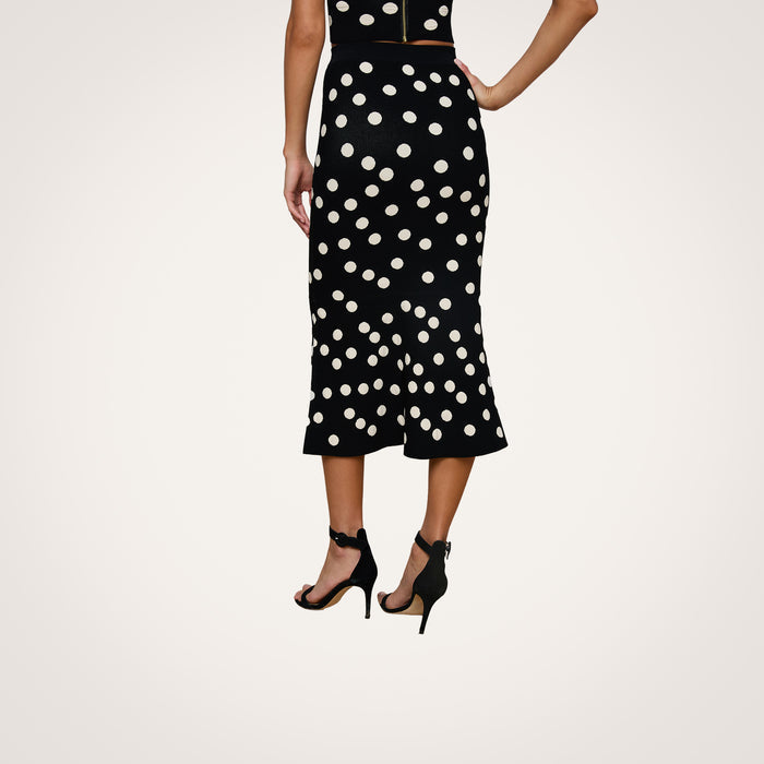A woman stands with one hand on her hip, wearing the Mae Knit Flare Skirt in black with white polka dots and black high-heeled sandals. The photo is taken from behind, showing only her lower back and legs.