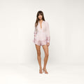 A woman wearing the Maela Short, a light pink long-sleeve romper with a floral print near the hem and cuffs and comfortable pull-on shorts, stands in heels against a plain white background.