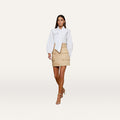 A woman walks forward wearing a white blouse with puffed sleeves, the Maelis Mini Skirt in beige featuring large pockets, and beige high-heeled sandals against a plain white background.