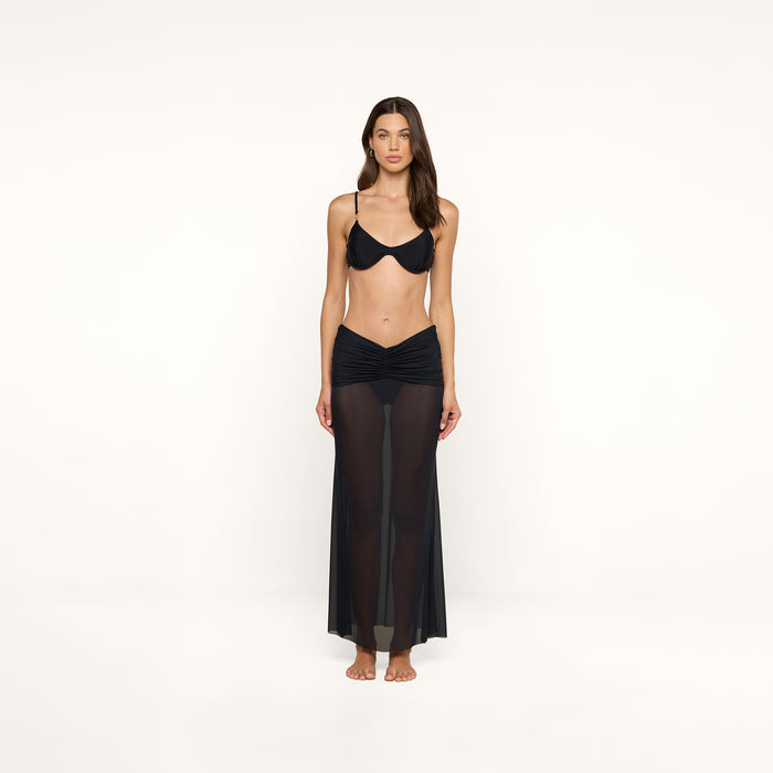 A woman stands barefoot against a plain white background, wearing a black bikini top and the Mahe Mesh Cover-Up Skirt.