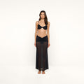 A woman stands barefoot against a plain white background, wearing a black bikini top and the Mahe Mesh Cover-Up Skirt.