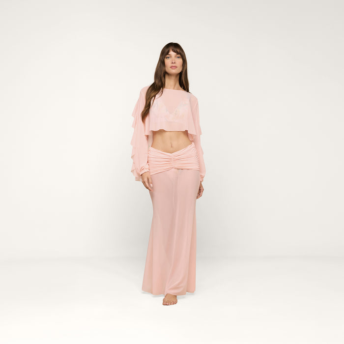A woman stands barefoot in a minimalist space, wearing the Mahe Mesh Cover-Up Skirt—a flowing, light pink, high-waisted mermaid skirt—paired with a matching long-sleeve crop top with ruffled sleeves.