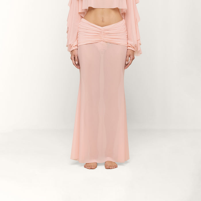 A person stands barefoot on a white background, wearing the light pink Mahe Mesh Cover-Up Skirt styled as a mermaid skirt and a matching long-sleeve top that reveals their midriff. The person’s head is not visible.