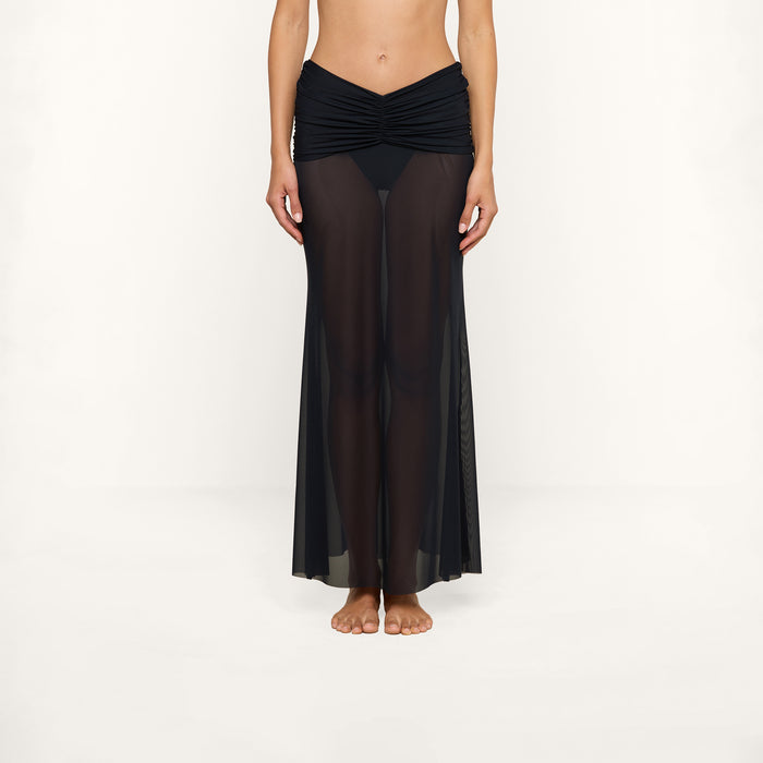 A person stands barefoot against a plain white background, wearing the Mahe Mesh Cover-Up Skirt—a black sheer mesh maxi skirt with a ruched waistband—over black underwear.