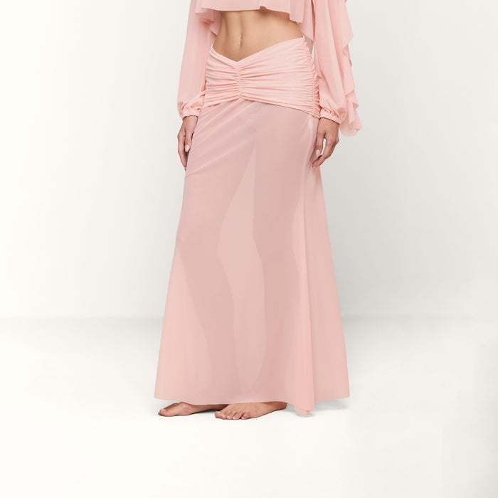 A person stands barefoot wearing a long, sheer, light pink Mahe Mesh Cover-Up Skirt with ruching at the waist and a matching cropped top. The background is plain white.