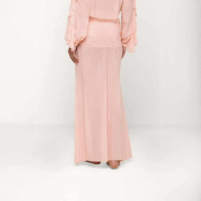 A person wearing a light pink, long-sleeved blouse and a matching pink Mahe Mesh Cover-Up Skirt stands barefoot against a plain white background, with their hands resting at their sides.