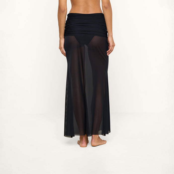 A woman stands barefoot facing away, wearing the Mahe Mesh Cover-Up Skirt—a sheer black maxi skirt with a high waistband—over a black bikini bottom. The background is plain and light.