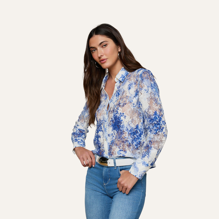 Woman with long brown hair wearing the Maia Lace Blouse in navy floral, styled with light blue jeans and a white belt, standing against a plain, light-colored background.