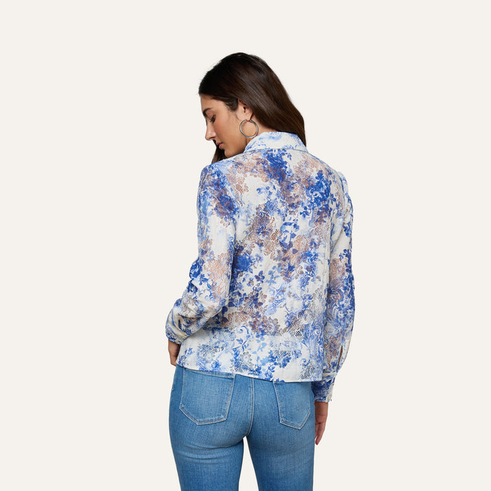 A woman with long brown hair stands with her back to the camera, wearing the Maia Lace Blouse in navy floral and blue jeans against a plain light background.