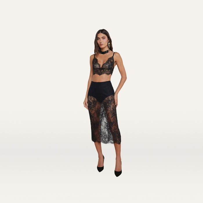 A woman wearing a black lace bralette, the Makena Lace Pencil Skirt with black shorts underneath, black heels, and statement earrings poses against a plain white background.