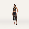 A woman wearing a black lace bralette, the Makena Lace Pencil Skirt with black shorts underneath, black heels, and statement earrings poses against a plain white background.