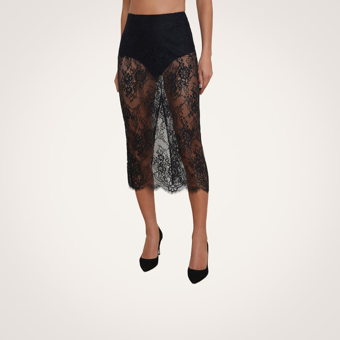A woman wearing the Makena Lace Pencil Skirt by L’AGENCE over black undergarments and black high-heeled shoes, standing against a light background.