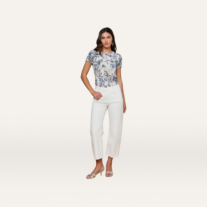 A woman stands against a plain white background, wearing a short-sleeve blue floral top, the Mallis Bow-Leg Jean in a high-rise wide-leg denim style, and open-toe heels. She has one hand on her hip and a neutral expression.