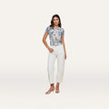 A woman stands against a plain white background, wearing a short-sleeve blue floral top, the Mallis Bow-Leg Jean in a high-rise wide-leg denim style, and open-toe heels. She has one hand on her hip and a neutral expression.