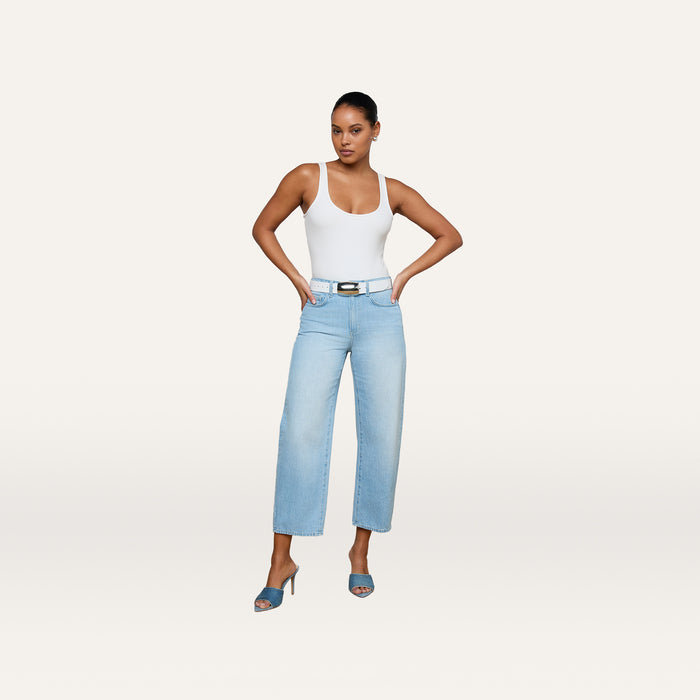 A woman in a white tank top, Mallis Bow-Leg Jean in light blue with a belt, and blue heeled sandals stands with hands on hips against a plain light background.