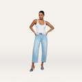 A woman in a white tank top, Mallis Bow-Leg Jean in light blue with a belt, and blue heeled sandals stands with hands on hips against a plain light background.