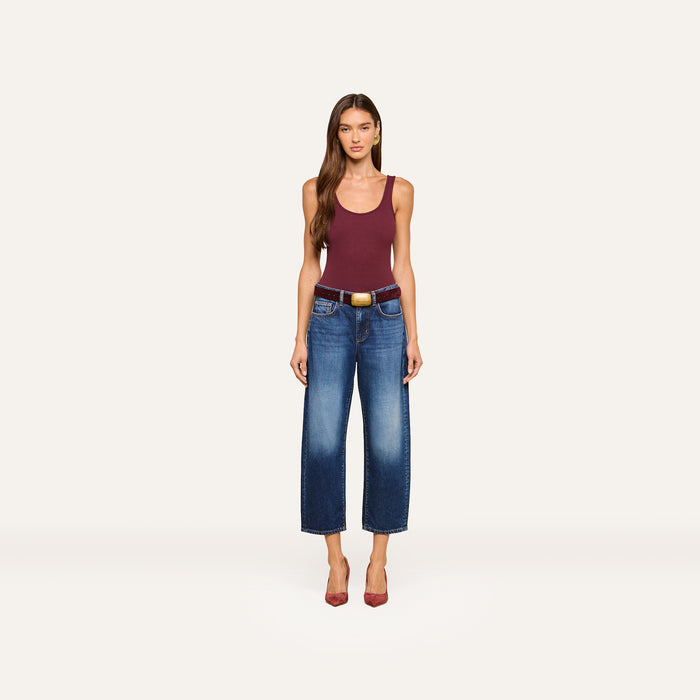 A woman stands facing forward, wearing a maroon tank top, the Mallis Bow-Leg Jean in indigo wash paired with a brown belt, and red heels, against a plain white background.