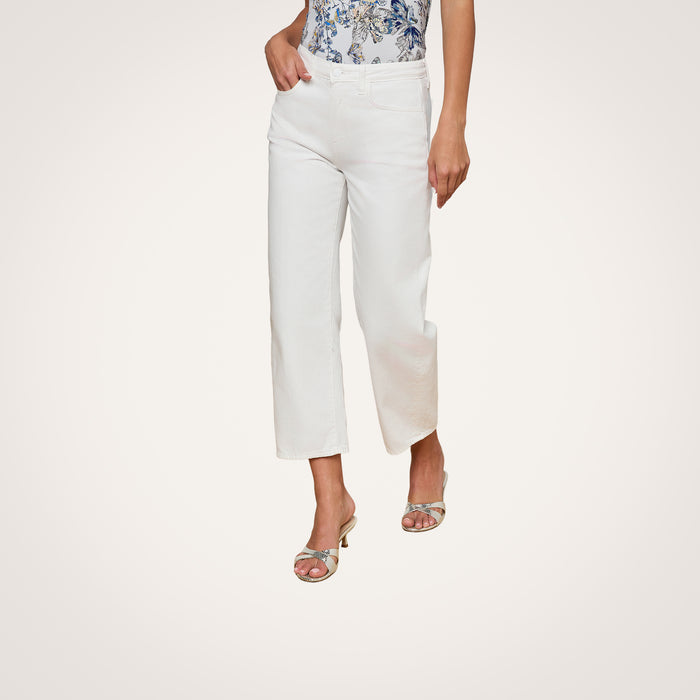 Wearing the Mallis Bow-Leg Jean in a high rise, white denim cropped style, paired with a patterned blue and white top and silver heeled sandals, a model’s lower body is shown standing against a plain white background.