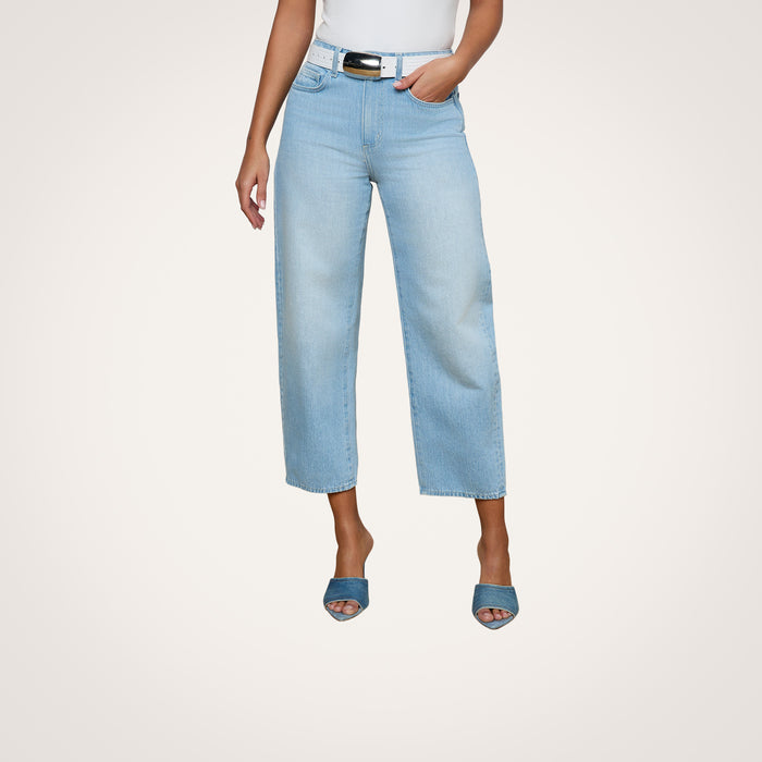 A person stands against a plain white background wearing the Mallis Bow-Leg Jean in light blue with a wide-leg, modern silhouette, paired with a white top and blue open-toe heels. The high-rise denim is styled fashionably, and the image is cropped above the shoulders.