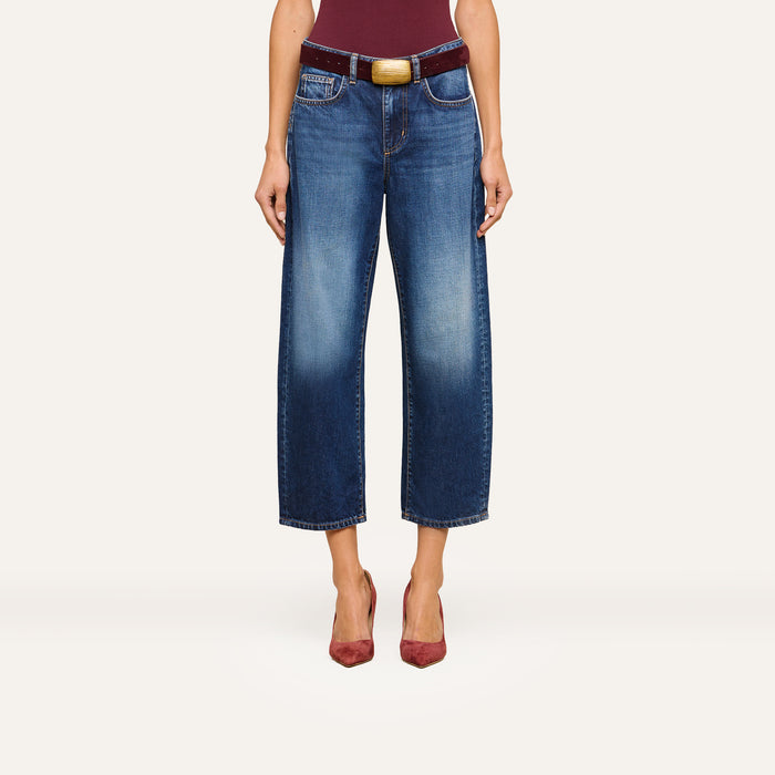 A person wearing the Mallis Bow-Leg Jean in indigo wash, paired with a burgundy belt featuring a large buckle and burgundy pointed-toe heels, stands against a plain background. The upper body is partially visible.