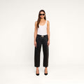 A woman stands against a plain white background, facing forward with her arms relaxed by her sides, wearing sunglasses, a white tank top, black heels, and the Mallis Coated Bow-Leg Jean in a high-rise, wide-leg silhouette.