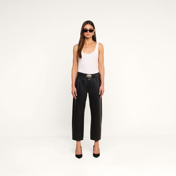 A woman stands against a plain white background, facing forward with her arms relaxed by her sides, wearing sunglasses, a white tank top, black heels, and the Mallis Coated Bow-Leg Jean in a high-rise, wide-leg silhouette.