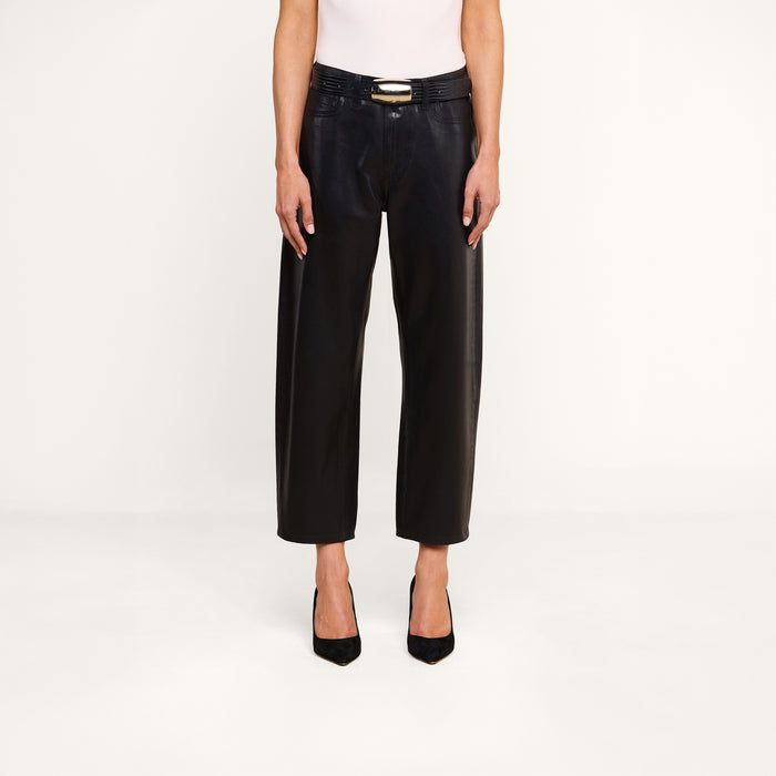 A person wearing the Mallis Coated Bow-Leg Jean—black coated denim pants in a high-rise, wide-leg silhouette—paired with a black belt, a white sleeveless top, and black heels stands against a plain white background. The person’s head is not visible in the image.