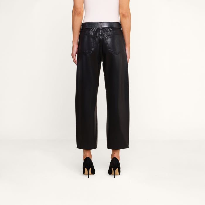 Model standing against a plain background, shown from the back, wearing the Mallis Coated Bow-Leg Jean in black with a high-rise and wide-leg silhouette, paired with black high-heeled shoes and a white sleeveless top.