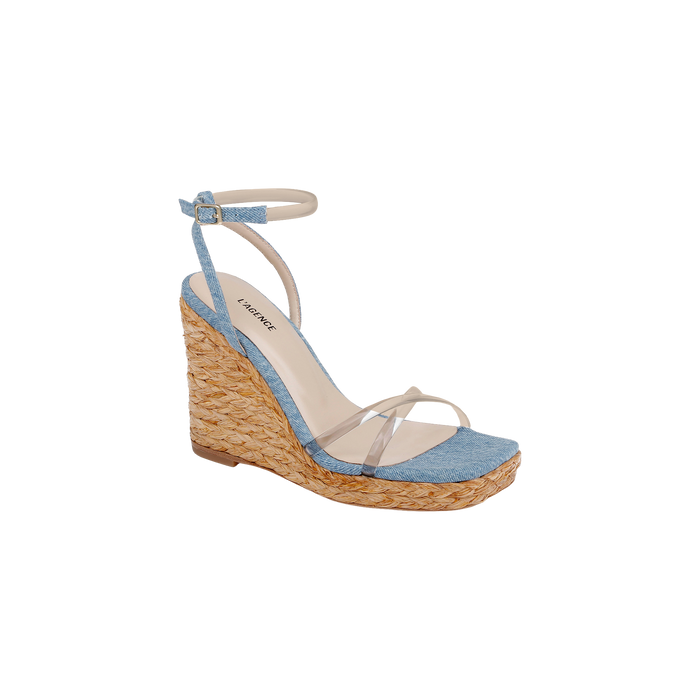 The Malone Wedge Espadrille is a bohemian-style wedge sandal showcasing a woven raffia heel, light blue fabric across the toe, clear straps over the foot, and a light ankle strap with buckle closure. Its cushioned beige insole provides comfort and is finished with a brand label.