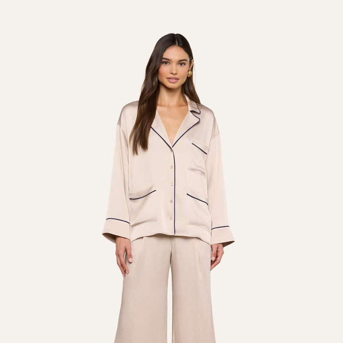 A woman stands facing forward, wearing the Mara Blouse — a cream-colored satin top with black piping and a relaxed fit button-up design. She pairs it with matching wide-leg pants, and stands against a plain light background.