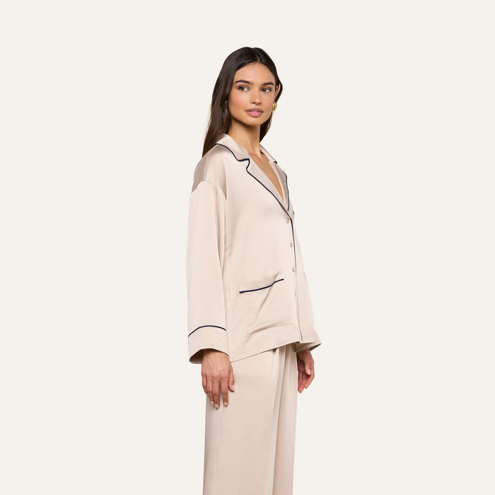 Model stands sideways, gazing slightly to the left, wearing the Mara Blouse in cream-colored satin with black trim and a relaxed fit. The plain, light background accentuates this elegant loungewear piece.