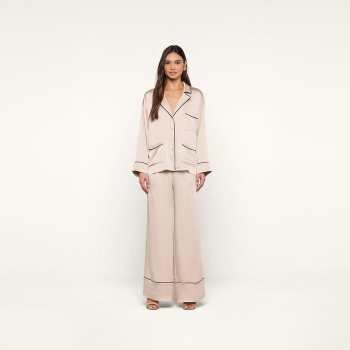 A woman stands facing forward, wearing the Mara Blouse in a relaxed fit light beige design with black piping details on the cuffs, pockets, and collar, against a plain white background.