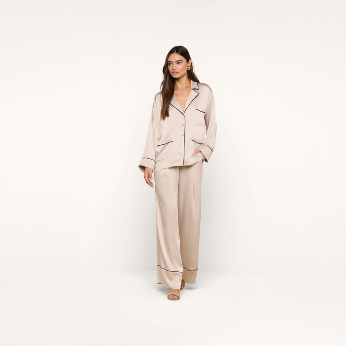 A woman stands against a plain white background, dressed in the Mara Blouse—a light beige satin top with dark trim—paired with open-toe sandals. The relaxed fit of the blouse enhances an effortless, comfortable style.