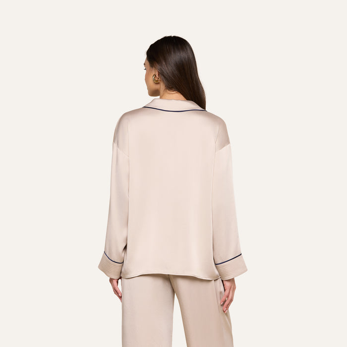 A woman with long brown hair is seen from behind, wearing the Mara Blouse—a relaxed fit, beige loungewear top with long sleeves and black piping details—standing against a plain light background.