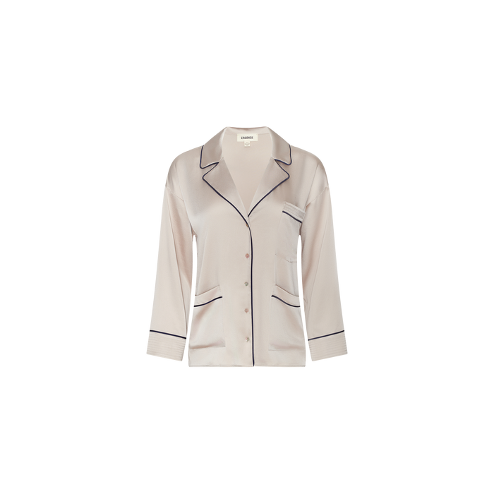 The Mara Blouse is a light pink satin pajama top featuring black piping, long sleeves, a notched collar, button front closure, and a single chest pocket. Displayed on a plain white background, this relaxed-fit blouse offers elevated comfort ideal for loungewear.