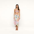 A woman stands barefoot against a plain white background, wearing a pastel floral bikini top and the Marena Sarong beach cover-up, featuring delicate fringe tassels.