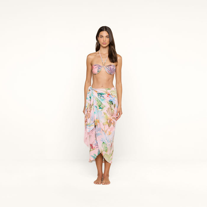 A woman stands barefoot against a plain white background, wearing a pastel floral bikini top and the Marena Sarong beach cover-up, featuring delicate fringe tassels.