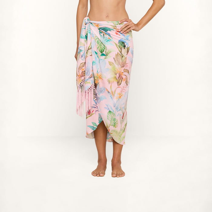 A person stands barefoot against a plain white background, wearing the Marena Sarong in a pastel tropical print, tied at the hip and styled as a beach cover-up.