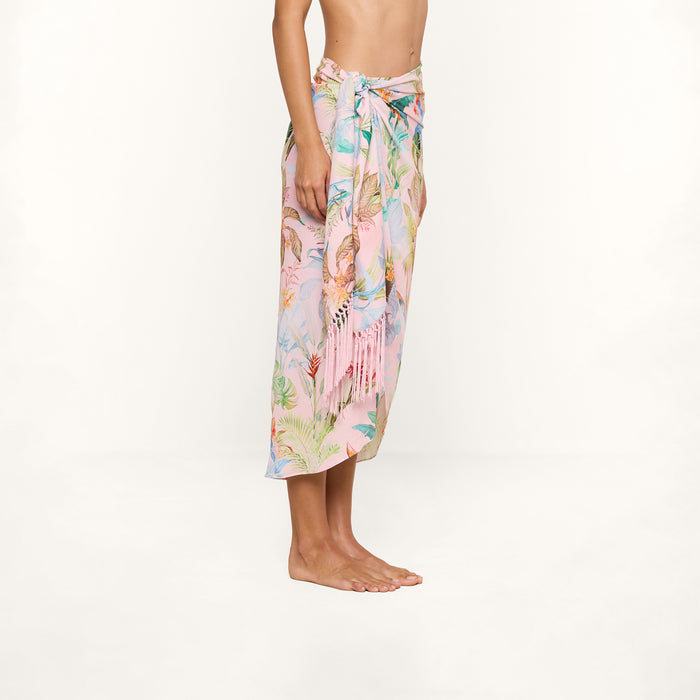 A person stands barefoot wearing the Marena Sarong—a vibrant, tropical-patterned skirt tied at the waist, perfect as a beach cover-up. The background is plain and white.