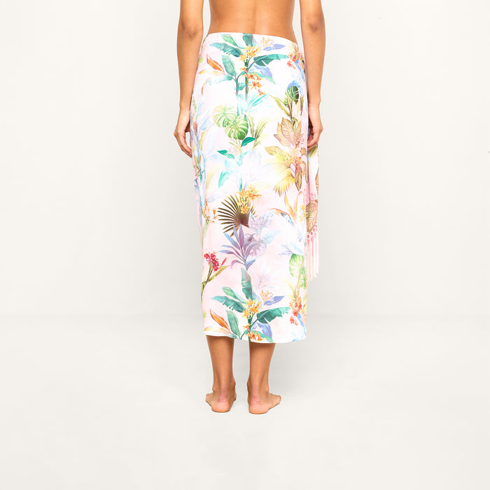 A person stands barefoot facing away, wearing the Marena Sarong—a long, light-colored wrap featuring a tropical floral pattern. Their upper body is uncovered, set against a plain, light background.