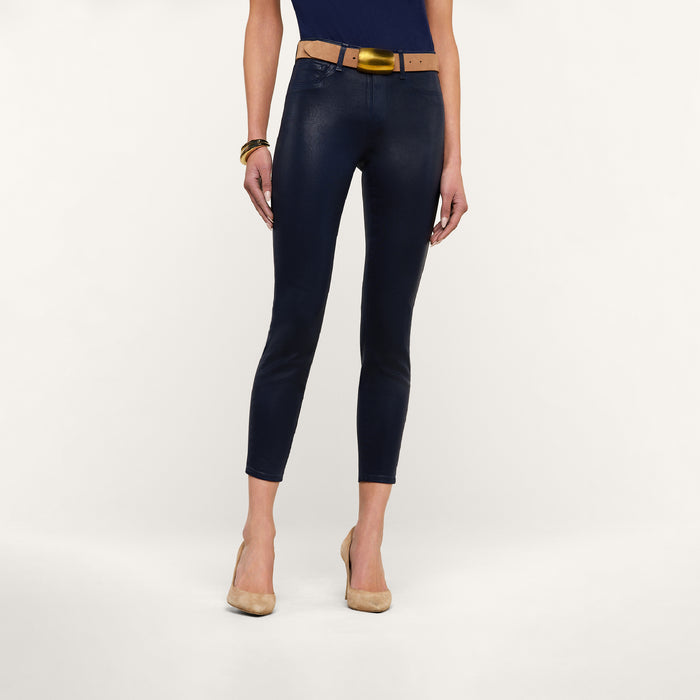 A woman stands wearing the Margot Coated Cropped Skinny Jean, featuring a contoured waistband and tapered leg in navy blue, paired with a tan belt, tan high heels, and a blue top. Her arms are relaxed at her sides with a brown bracelet on her right wrist. The background is plain white.