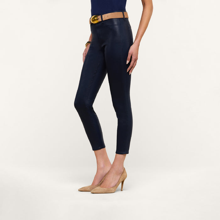 A woman stands against a white background wearing the Margot Coated Cropped Skinny Jean in navy with a tapered leg, styled with a tan belt featuring a gold buckle, a navy top, and tan high heels.